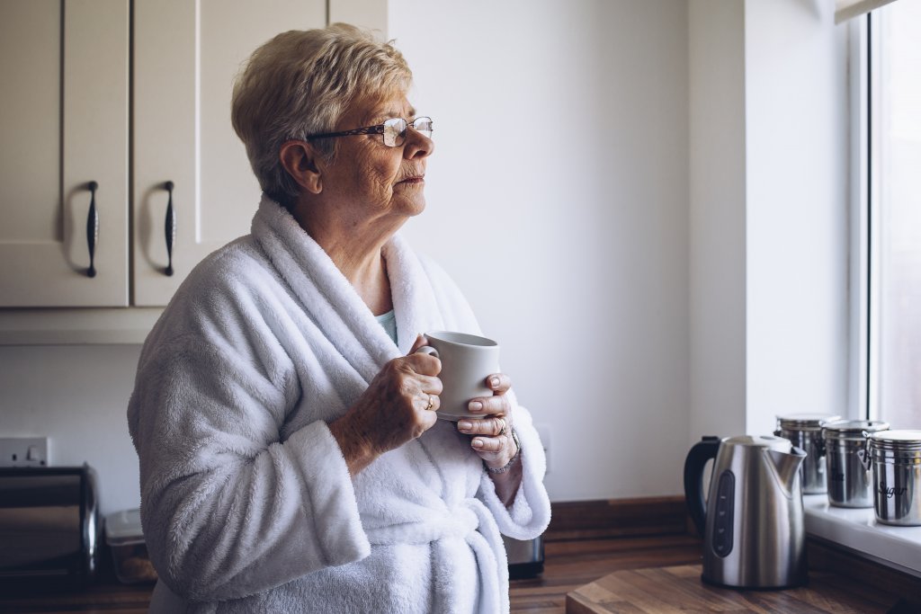 Elderly woman living alone and looking out of the window with tea in hand.
